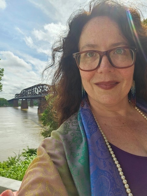 Monica Clark-Robinson, a white woman with curly dark hair, brown glasses and a rainbow colored scarf, smiles in front of the Mississippi River.